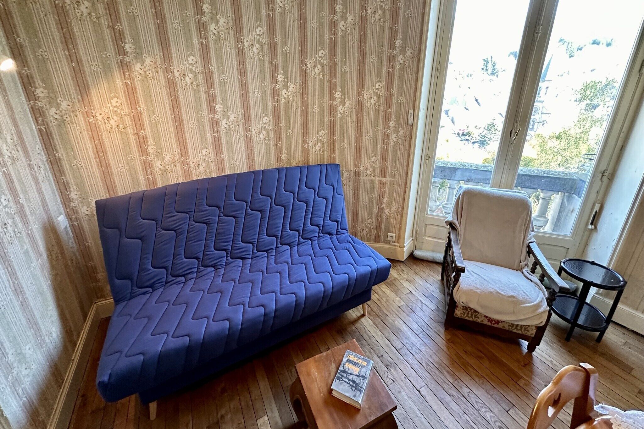 Une pièce avec un canapé bleu, une chaise en bois, une petite table sur laquelle se trouve un livre, et une vue par la fenêtre.