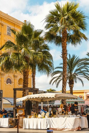 Un marché de rue avec des étals où sont exposés divers articles, des palmiers et, au fond, un bâtiment jaune.