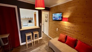 Una camera accogliente con un letto, una TV fissata al muro, una panca in legno e una piccola area cucina visibile attraverso una porta aperta.