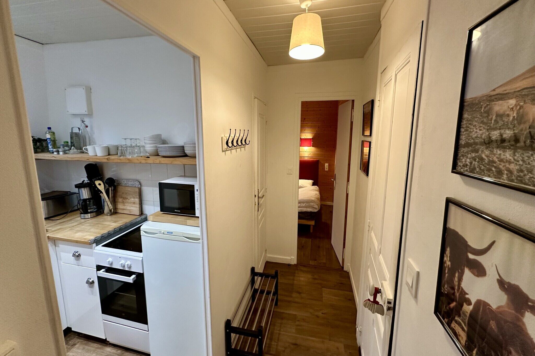 A narrow hallway with a kitchen area on the left, featuring a stove, oven, and various kitchen items. A bench runs along the hallway, and there are framed pictures on the walls.