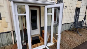 Eine weiße Tür mit Glasfeldern, eine schwarze Fußmatte und ein Metallstuhl vor einem Gebäude mit beige gefärbten Backsteinwänden.