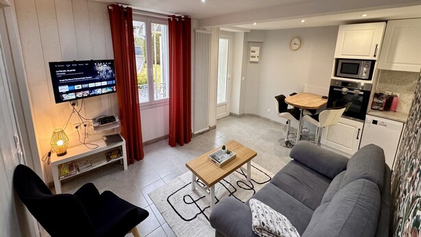 Ein modernes Wohnzimmer mit grauem Sofa, einem Holztisch, einem an der Wand befestigten Fernseher und einer Küchenzeile mit weißen Schränken und einer Mikrowelle.