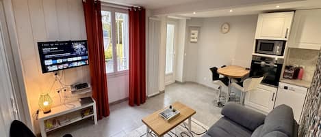 A modern living room with a grey sofa, a wooden coffee table, a TV mounted on the wall, and a kitchen area with white cabinets and a microwave.