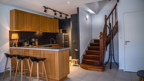 A modern kitchen with wooden cabinets, a central island with bar stools, a built-in microwave, and a stainless steel refrigerator. A wooden staircase with a railing leads to a door. - Estate with Panoramic Sancy Views (Mont-Dore)