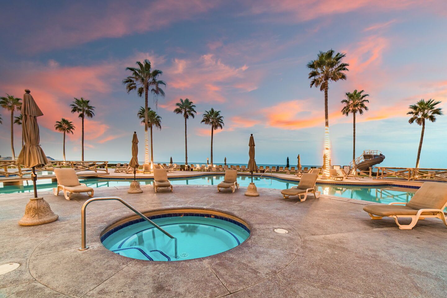A pool area with lounge chairs and umbrellas, surrounded by palm trees, during sunset.