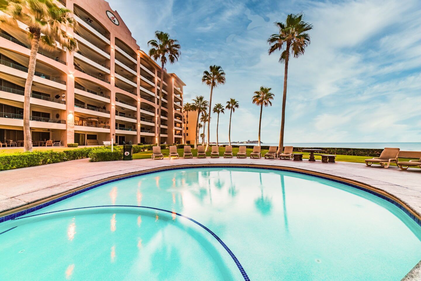 Una piscina con sillas de descanso y palmeras, rodeada por un edificio de hotel con varios pisos y balcones.