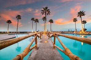 Vista de la piscina con muelle de madera, palmeras y cielo al atardecer.