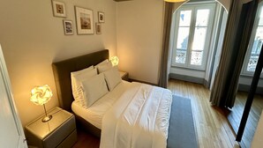 A neatly made bed with white linens, a bedside lamp, and a nightstand. There are framed pictures on the wall above the bed. - Quiet corner near the mountain road (Mont-Dore)