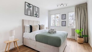 A modern bedroom with a large bed, a bedside table with a lamp, a potted plant, and artwork on the walls. - Cozy 1BR Near Hyde Park & Notting Hill (London)