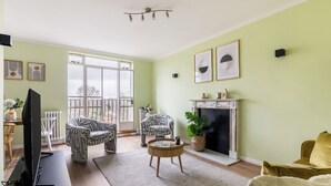 A living room with a fireplace, two patterned armchairs, a yellow sofa, and a flat-screen TV on a stand. - Cozy 1BR Near Hyde Park & Notting Hill (London)
