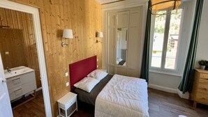 A bedroom with a wooden wall, a bed with white bedding, a nightstand, a small bedside table, a mirror, and a window with curtains. - Quiet corner near the mountain road (Mont-Dore)