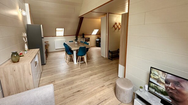 Cocina compacta y moderna con suelo de madera, zona de comedor con sillas azules y un televisor sobre un mueble.