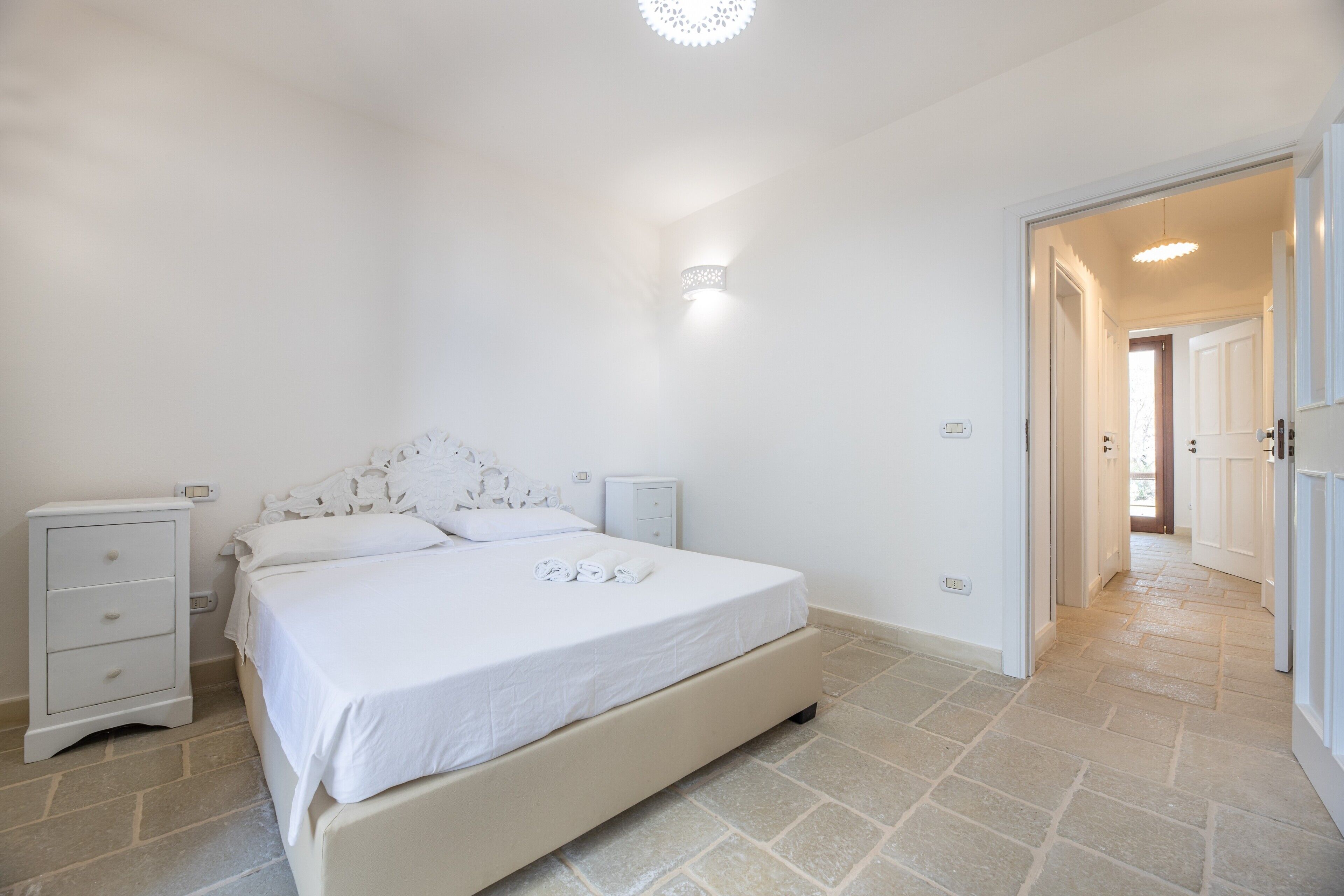 A bedroom with a bed, bedside tables, a nightstand, a wall-mounted light, and a door leading to another room.
