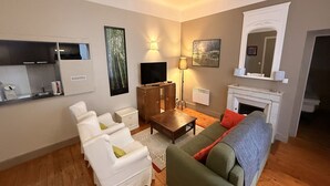 Un salon comprenant un canapé, des fauteuils, un téléviseur, une cheminée et un coin cuisine.