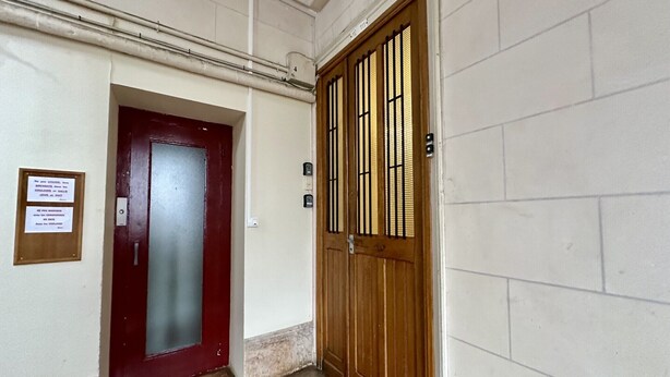 Un couloir avec une porte rouge et une porte en bois ornée de barreaux verticaux. Il y a un panneau sur le mur et un interrupteur près de la porte en bois.