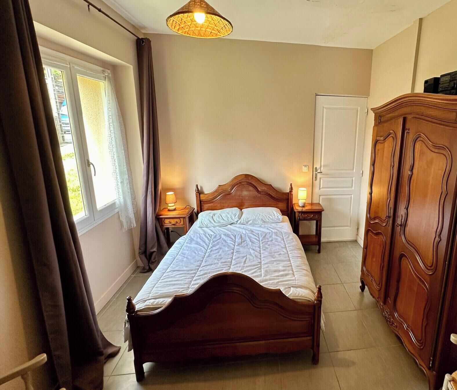 A bedroom with a wooden bed, two bedside tables with lamps, a wardrobe, and a window with curtains.
