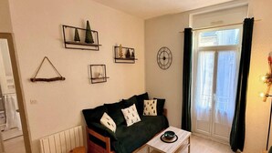 Ein gemütliches Wohnzimmer mit einem dunkelgrünen Sofa, einem kleinen Holztisch und einer Heizung. An den Wänden befinden sich Dekorationen, darunter eine Uhr und gerahmte Bilder.