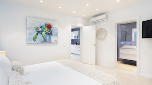Ein Schlafzimmer mit einem großen Bett, einem Gemälde einer Meeresschildkröte, einer Klimaanlage und Blick ins Bad mit einer blauen Fliesenwand.