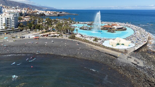 Ein Küstenort mit einem breiten Sandstrand, einem zentral gelegenen Pool und einer felsigen Küstenlinie.