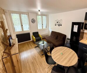 A cozy living room with a brown sofa, a yellow armchair, a glass coffee table, and a wooden dining table with chairs. There are two windows with curtains, a wall clock, and a refrigerator with a note attached. - Family Hub with Modern Amenities (Mont-Dore)