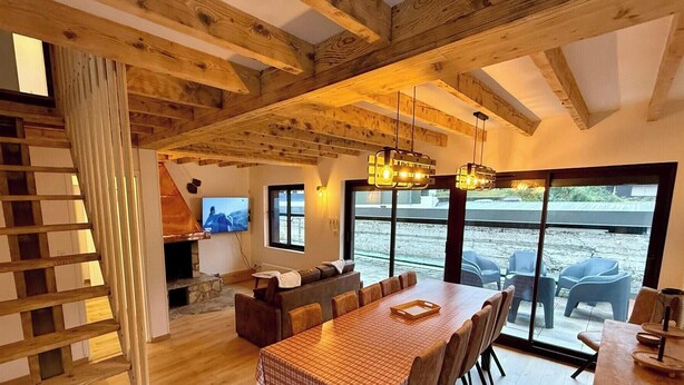 Een ruime woonkamer met houten plafond, een open haard, een televisie en een groot raam met uitzicht op een prachtig buitengebied.