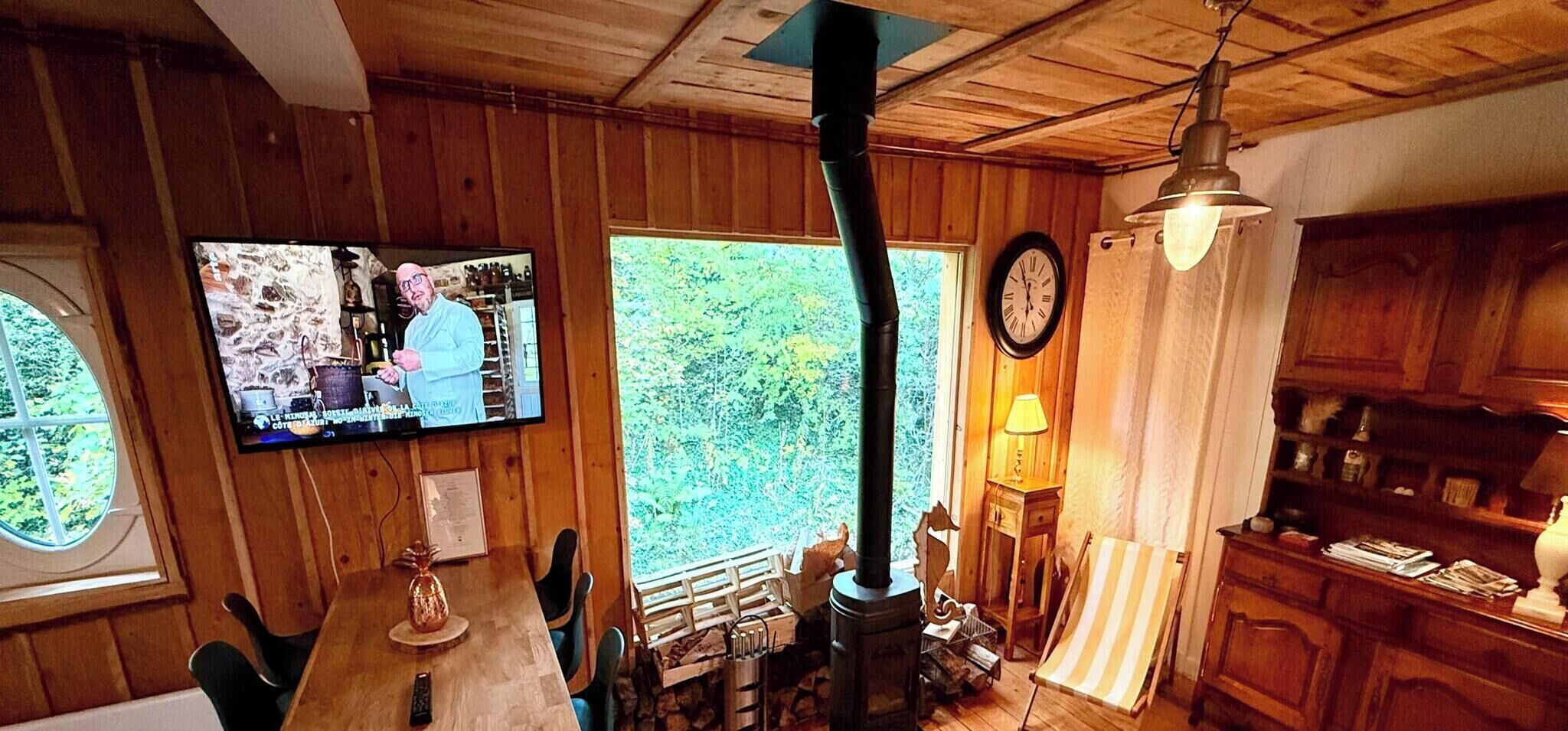 Et træpanelt interiør med et spisebord, stole, et fjernsyn, et ur og et vindue med udsigt til grønne omgivelser.