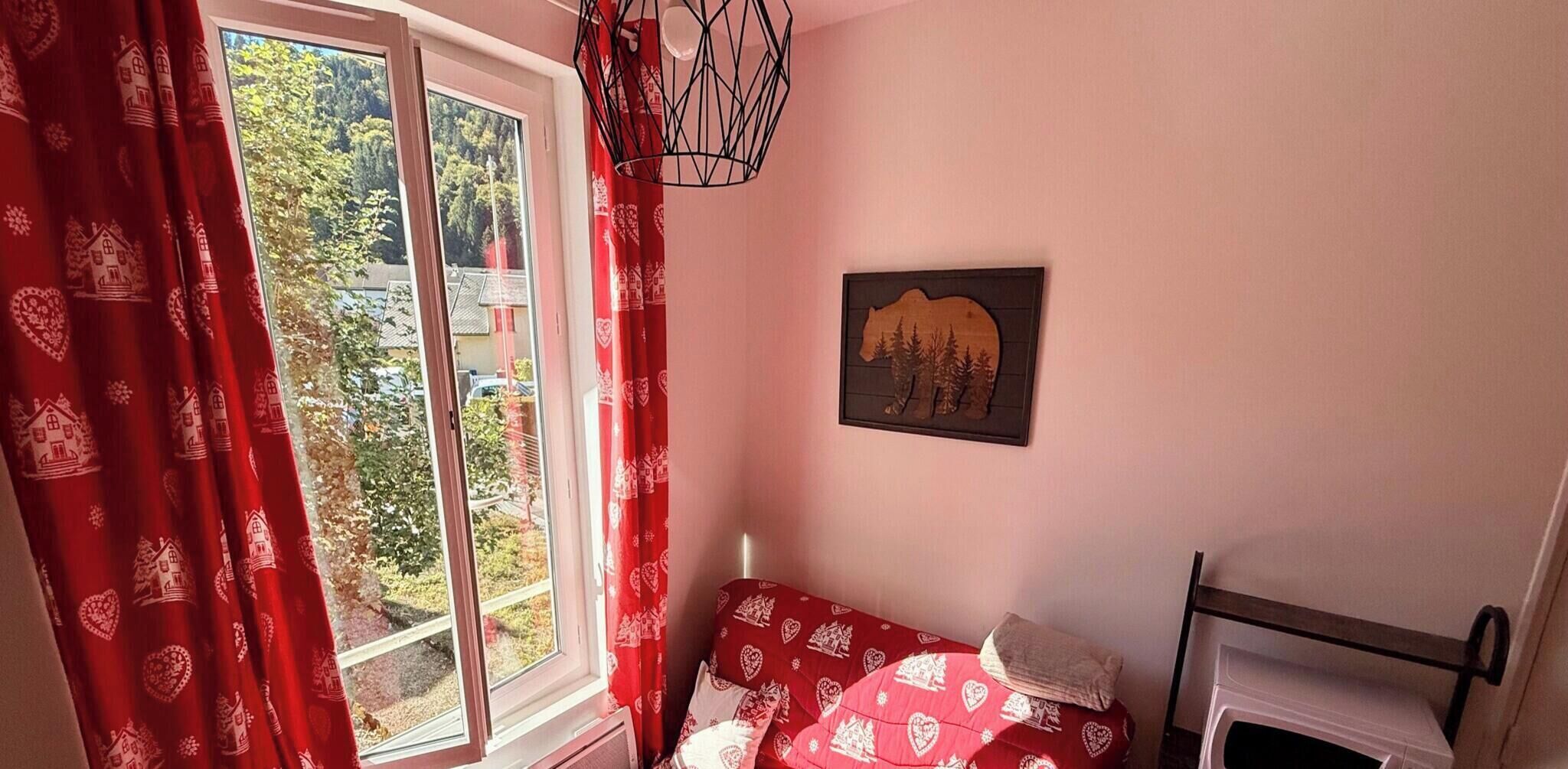 A bedroom with a bed, a window with red curtains, a wall-mounted light fixture, and a framed picture of a bear.