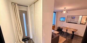 A modern living room with a dining area, a TV mounted on the wall, and a large window. - Modern nook by the central way (Mont-Dore)