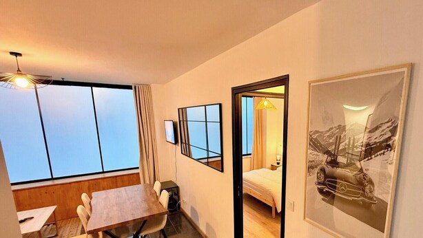 A hotel room with a large window, a bed, a desk, and a chair. There is a framed picture on the wall and a lamp on the desk. - Modern nook by the central way (Mont-Dore)