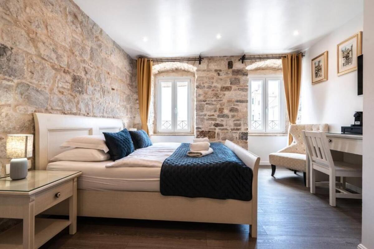A bedroom with a stone wall, a bed with white linens, a desk, and a chair.