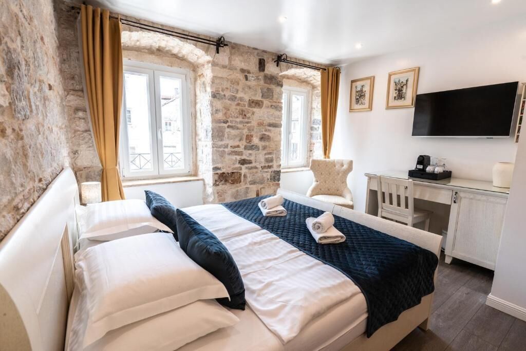 Habitación de hotel con pared de piedra, cama con ropa de cama blanca y azul oscuro, escritorio con televisor y ventana con cortinas.