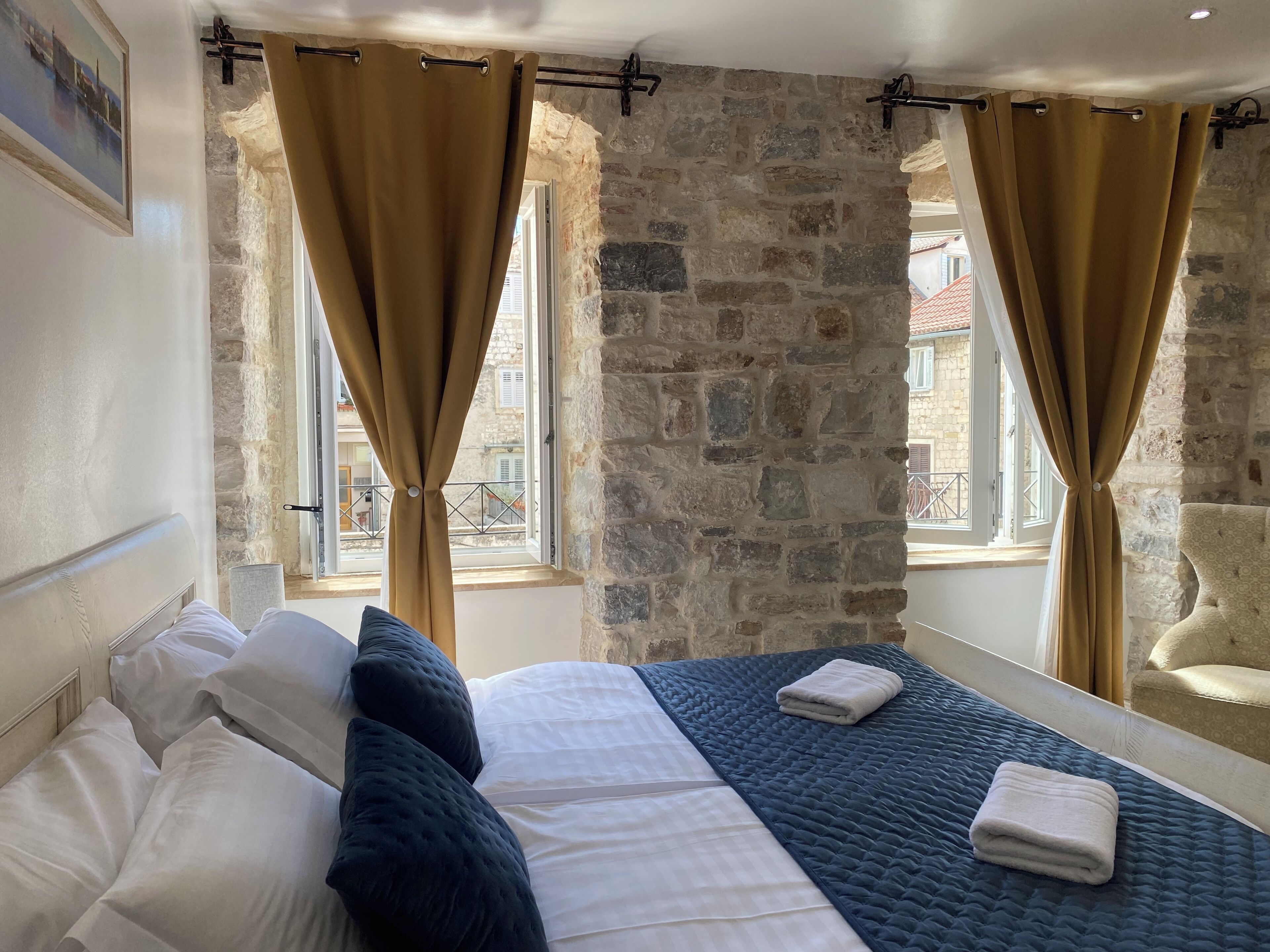 A bedroom with a stone wall, a bed with white and dark blue bedding, and two windows with curtains.
