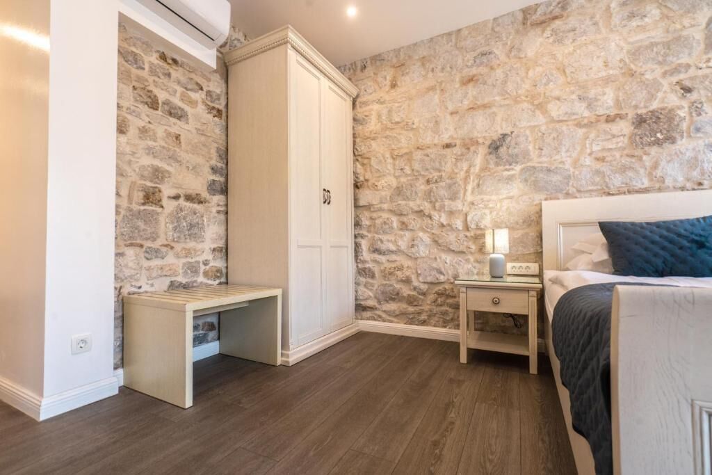 A bedroom with a stone wall, a wardrobe, a bedside table with a lamp, and a bed with a blue pillow.