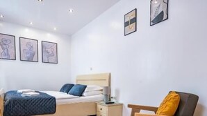 Une chambre à coucher avec un lit, une table de chevet, une lampe, une chaise et une œuvre d’art accrochée au mur.