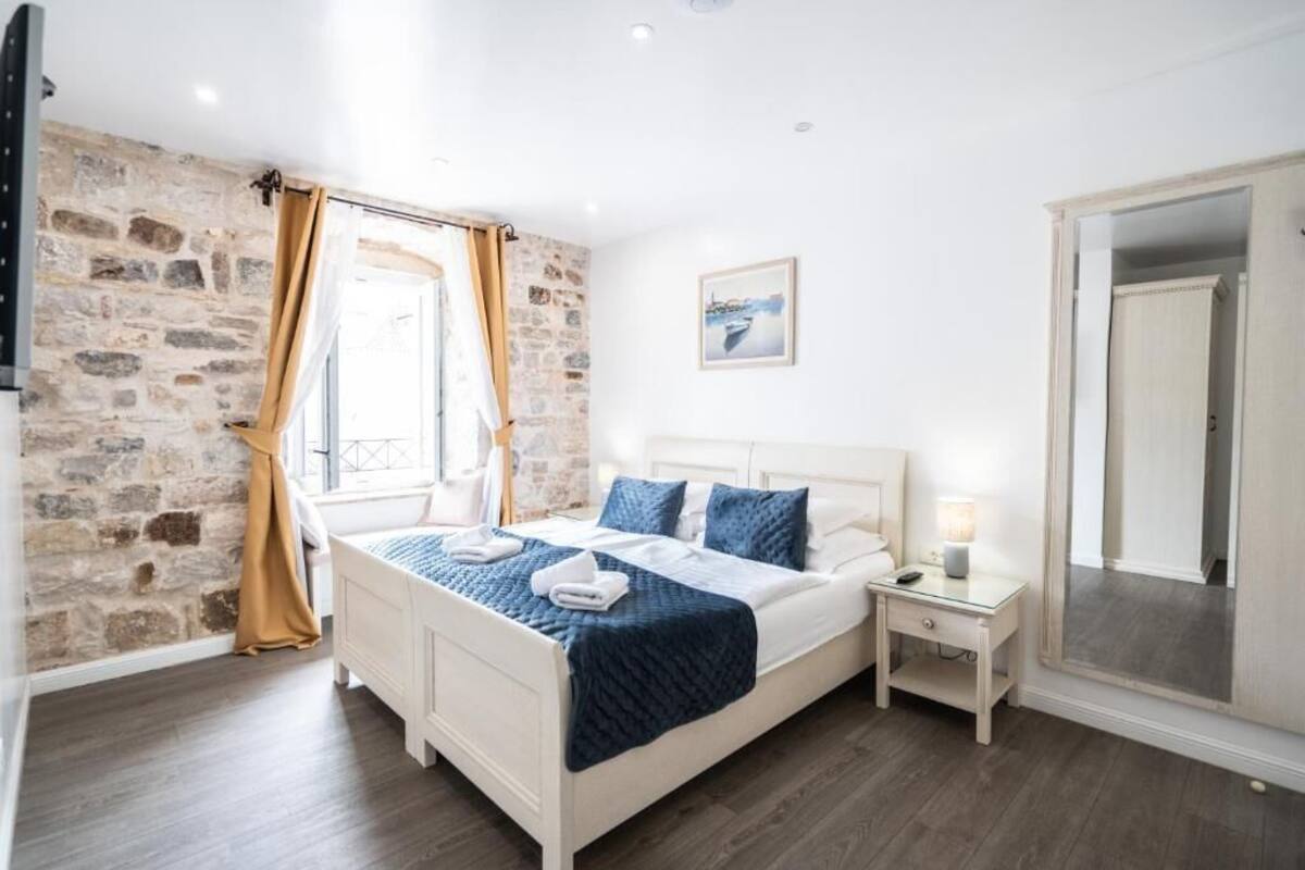 A bedroom with a stone wall, a bed with blue bedding, a bedside table with a lamp, and a large mirror.