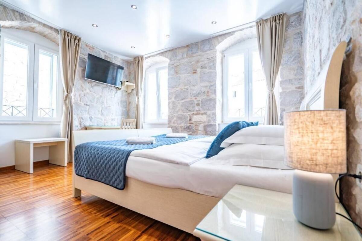 A bedroom with a stone wall, a bed with a blue blanket, a bedside table, a lamp, and a TV mounted on the wall.