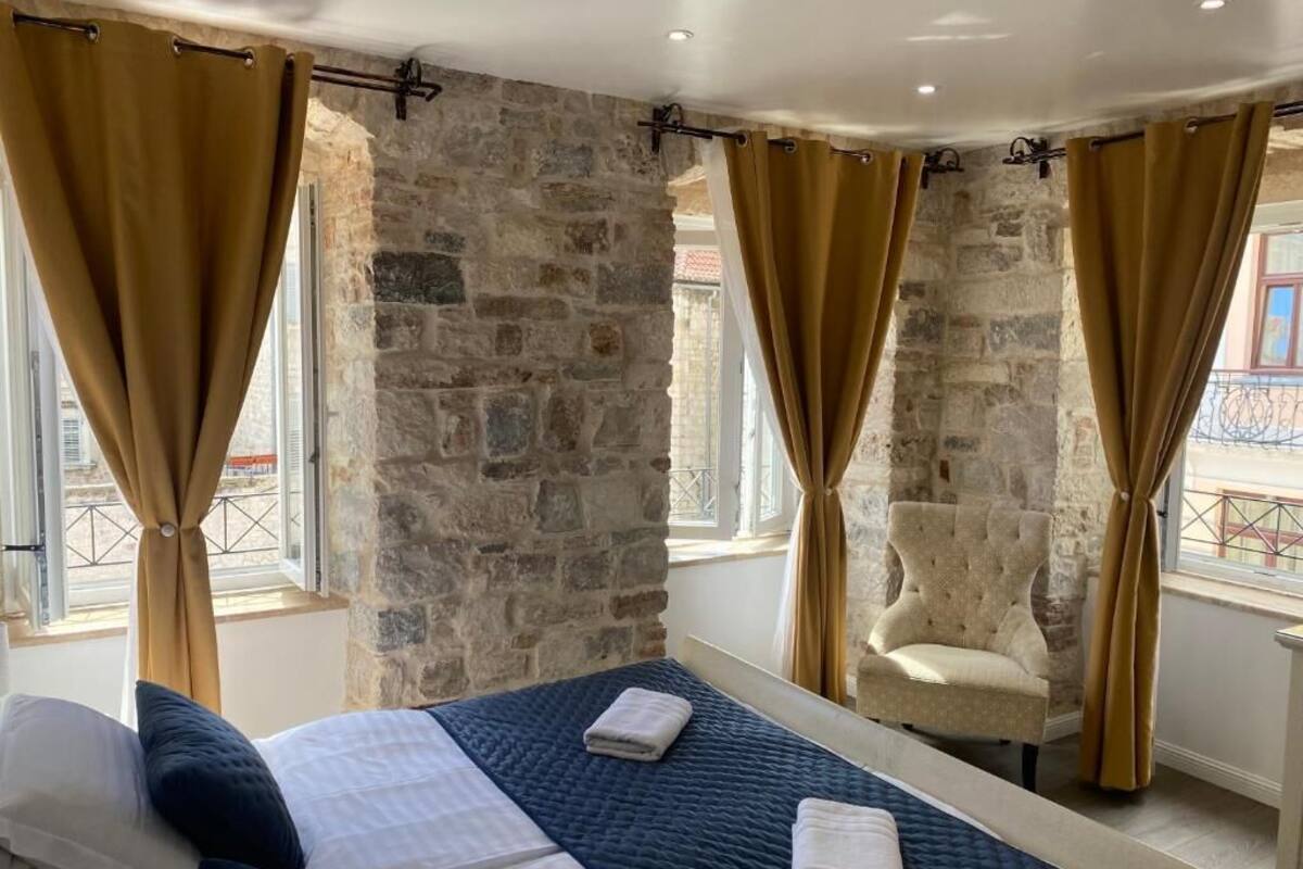 A room with a stone wall, two beds with blue and white bedding, a chair, and curtains.
