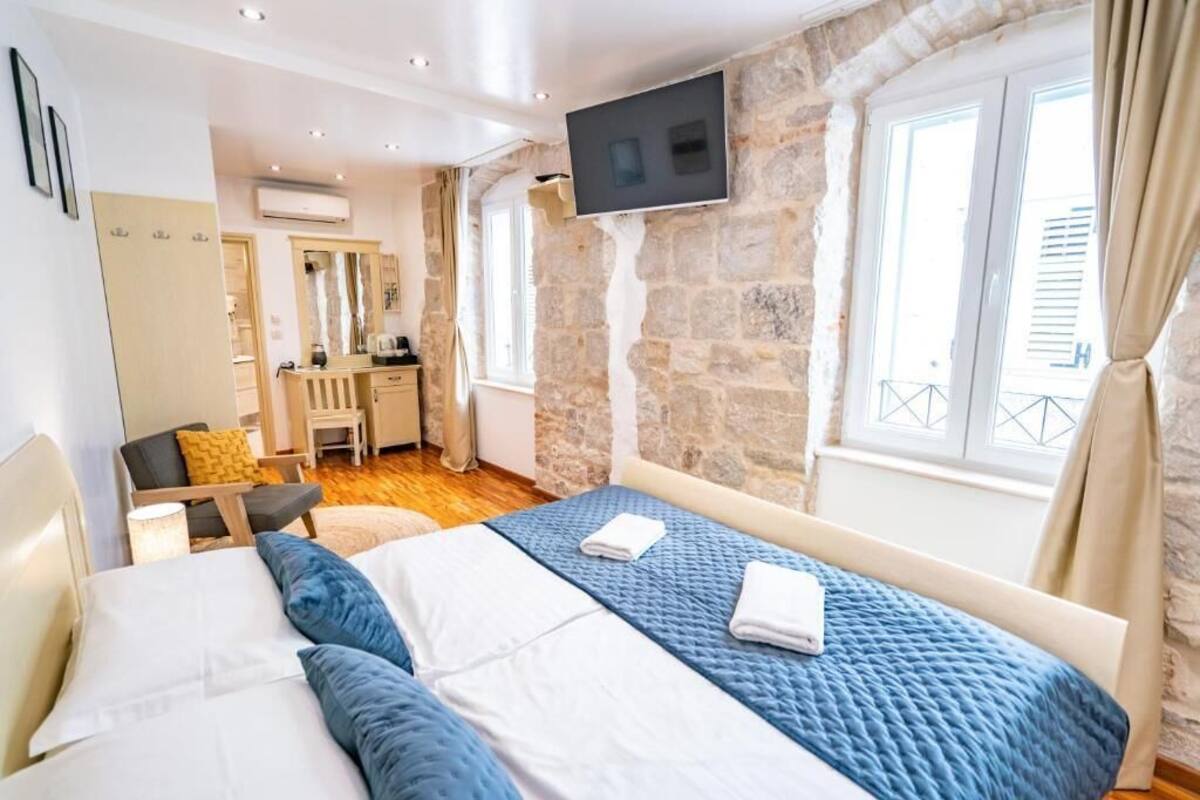 A bedroom with a bed, a desk, a chair, a TV mounted on a stone wall, and a window with curtains.