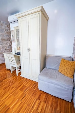 Une pièce avec un plancher en bois, une armoire blanche, un canapé gris agrémenté d’un coussin à motifs et un bureau blanc avec une chaise.