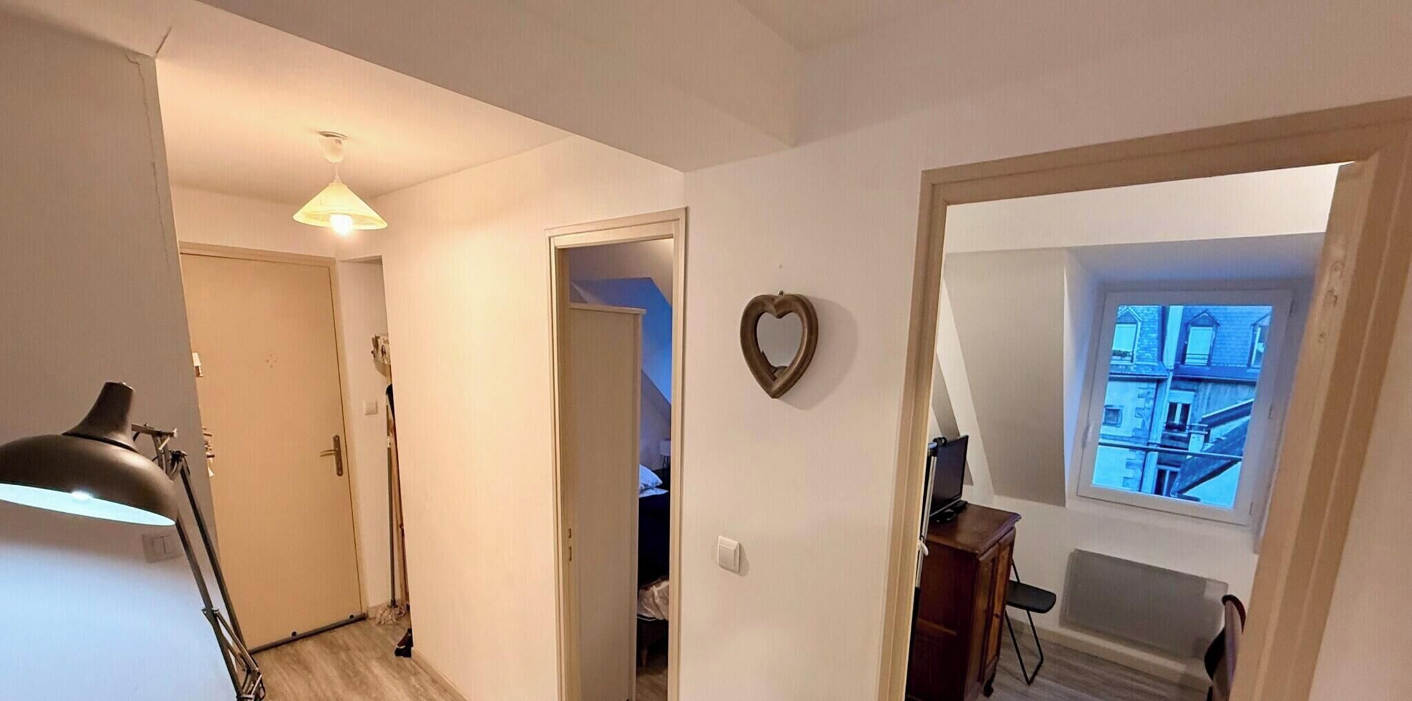 A narrow hallway with a ceiling light, a heart-shaped decoration on the wall, and a window with a view of buildings.