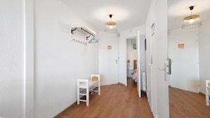 Passadís amb parets blanques, tarima de fusta, un penjallot, una taula petita amb una cistella i una porta que dona a una altra habitació.