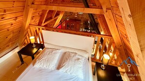Quarto de cabana de madeira com cama, mesas de cabeceira e vista de um edifício de pedra pela janela.