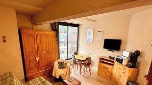Ein gemütliches Zimmer mit einem Holzschrank, einem Esstisch mit Stühlen, einem Fernseher und einem Fenster mit Blick auf Bäume.