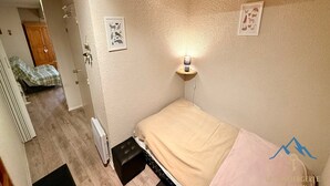 Ein kleines, gemütliches Schlafzimmer mit einem Einzelbett, einem Nachttisch mit Lampe und einem Heizkörper. Eine Tür führt in einen anderen Raum, und an der Wand hängt ein gerahmtes Bild.