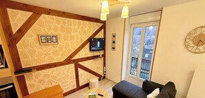 A cozy living room with exposed wooden beams, a stone wall, a flat-screen TV, a balcony with a view, and a round clock on the wall. - Grand hub near the village (Mont-Dore)