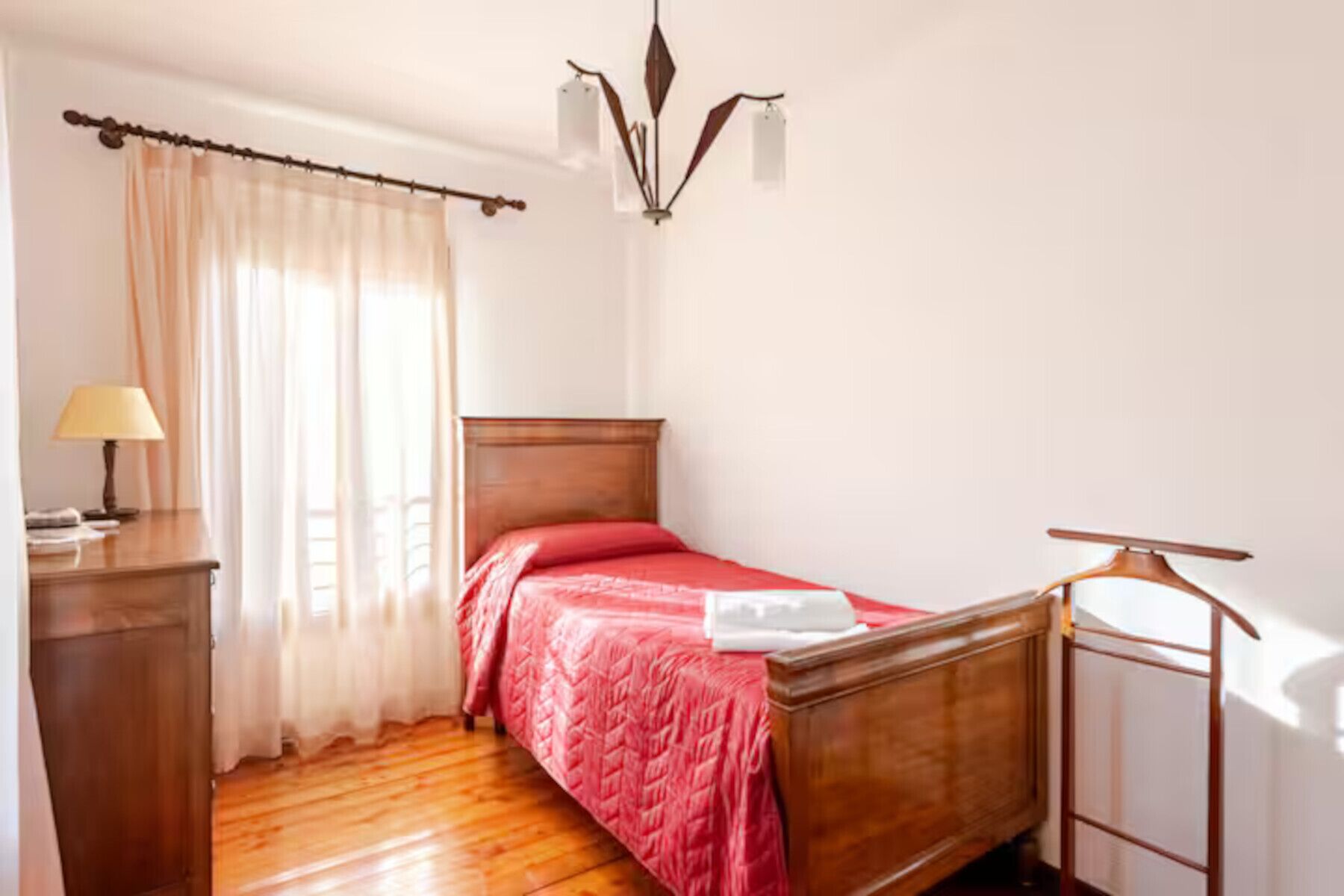 Een slaapkamer met een houten bed, een nachtkastje met een lamp en een raam met dunne gordijnen.
