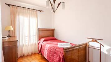 Een slaapkamer met een houten bed, een nachtkastje met een lamp en een raam met dunne gordijnen.