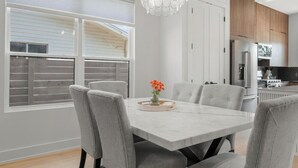 Uma sala de jantar moderna com uma mesa branca e cadeiras cinzentas, um candelabro e uma janela com persianas.