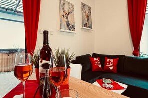 Ein Zimmer mit einem Holztisch, zwei Weingläsern, einer Weinflasche und einer Topfpflanze. Es gibt eine schwarze Couch mit roten Kissen sowie zwei Wandbilder von Rehen.