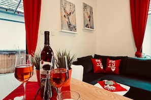 A room with a wooden table, two wine glasses, a wine bottle, and a potted plant. There is a black couch with red cushions and two wall paintings of deer. - Holiday home with jacuzzi, for 6 people (Rozewie)